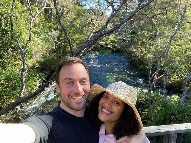 Cynthia Addai-Robinson is wearing a pink hoodie and cream color hat, whereas Thomas is wearing a black t-shirt.